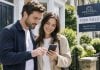 Prospective homebuyers viewing information on a smartphone while standing outside a property with a visible estate agent's for sale board.